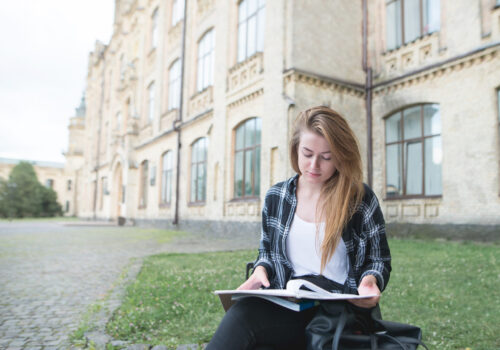 Concentrated beautiful girl student sitting on a bench on college campus and reading. Beautiful girl in casual clothes outdoors studying at university. Student Concept